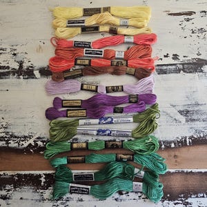 May include: A collection of twelve skeins of six-strand embroidery floss arranged in a vertical column. The colors include shades of yellow, coral, orange, brown, lavender, purple, and various tones of green, each featuring a paper label.