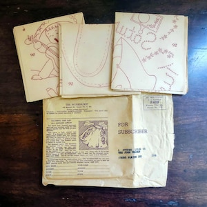 以下が含まれることがあります： ヴィンテージの裁縫パターン小冊子と封筒。小冊子にはイラストと番号90、91、92が描かれています。「The Workbasket」と「For Subscriber」と書かれた封筒。濃い木製の表面に置かれています。
