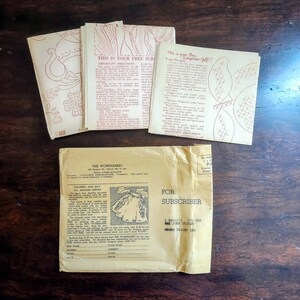 May include: Vintage sewing patterns and instructions. Several booklets with red illustrations and text are stacked on a wooden surface. An envelope with text "THE WORKBASKET" and "FOR SUBSCRIBER" is also present.