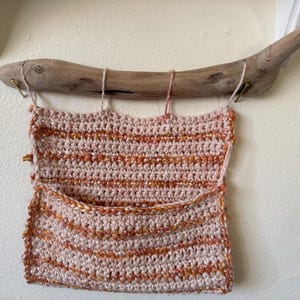 May include: A handmade crochet wall pocket in shades of pink and orange, suspended from a piece of driftwood. The pocket features a textured, striped design and a small pocket at the bottom, perfect for storing small items. The piece is hanging on a white wall.