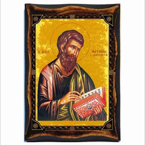 Puede incluir: Un icono religioso de San Mateo, el evangelista, sosteniendo un libro. El icono está pintado en un estilo tradicional con detalles dorados y un marco de madera oscura.