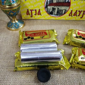 May include: A box of charcoal briquettes for incense burning. The box is yellow with black text that reads "Karvounákia Agia Lavra" and a picture of a church. The box is sitting on a burlap surface with two silver foil-wrapped charcoal briquettes and a black charcoal disc.