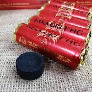 May include: A close-up of a stack of red and gold quick-lighting charcoal briquettes. The briquettes are wrapped in foil and have the text "MAAGRITHC" printed on them. The briquettes are 27 mm in diameter. A single black charcoal disc is in the foreground.