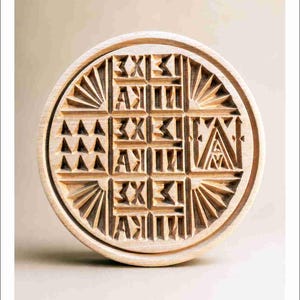 Könnte beinhalten: Ein kreisförmiger Stempel aus hellem Holz mit komplexen, geschnitzten geometrischen Mustern, darunter Reihen von Dreiecken, Sonnenstrahlen und wiederholtem Text "ΣΧΙ" und "ΛΙΚΑ", der in einem Gitterdesign auf der flachen Oberfläche angeordnet ist.
