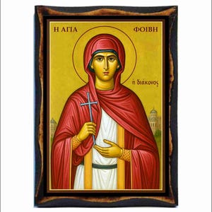 May include: Icon of a saint in a red robe and hood, holding a cross. The icon has a gold halo and is set in a dark wooden frame. The background is gold with Greek text.