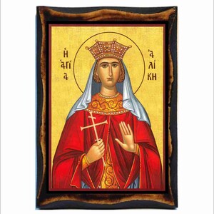 Puede incluir: Un icono religioso que representa a una mujer con una corona y una túnica roja, sosteniendo una cruz. El icono está enmarcado en un marco de madera oscura. El texto en el icono dice "Η άγια" y "άι κη"