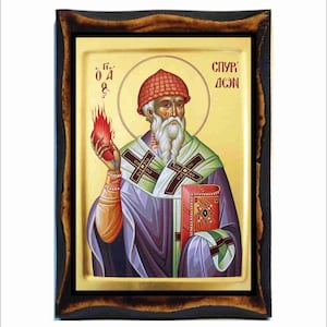 Puede incluir: Un icono religioso de un hombre barbudo con un sombrero rojo, sosteniendo un corazón en llamas en su mano izquierda y un libro en su mano derecha. El icono está enmarcado en un marco de madera oscura y tiene un fondo dorado. El texto "СВ. АСОН" está escrito en la esquina superior derecha del icono.