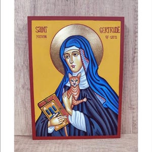 Puede incluir: Un icono de madera pintado a mano de Santa Gertrudis de Nivelles, la santa patrona de los gatos. El icono representa a Santa Gertrudis sosteniendo un libro y un gato jengibre. El fondo es de color amarillo dorado con un halo dorado alrededor de la cabeza de Santa Gertrudis. El icono está enmarcado en rojo.