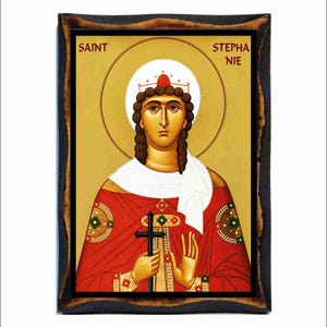 May include: A rectangular wooden plaque featuring a religious icon of Saint Stephanie. The figure wears a red robe with gold accents, a white head covering, and a crown, holding a black cross. The text SAINT STEPHANIE is printed in red.