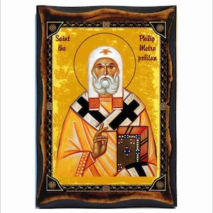 May include: A religious icon of Saint Philip the Metropolitan, painted in a traditional style on a wooden panel. The icon features a bearded man in a yellow robe and black vestments, holding a book. The background is a gold colour with a distressed texture.