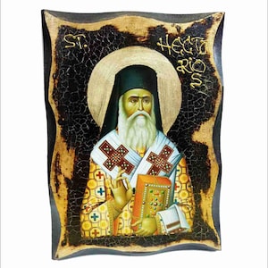 May include: A hand-painted icon of a saint holding a book. The icon is framed in a dark wood frame with a distressed finish. The background is black with a gold halo around the saint's head. The text "ST. HEORIOS" is written in gold lettering at the top of the icon.