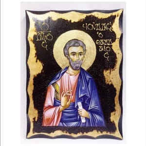 Op de afbeelding: Een handgeschilderd icoon van een man met een baard en een halo, die een blauwe robe draagt en een rol papier vasthoudt. Het icoon is ingelijst in een donkerhouten lijst met gouden accenten. De tekst "ΙΩΑΝΝΗΣ Ο ΘΕΟΛΟΓΟΣ" is in gouden letters op de lijst geschreven.