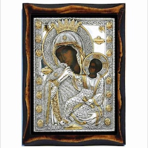 Può includere: Un'icona in argento e oro della Vergine Maria e il bambino Gesù. L'icona è incorniciata in una cornice di legno scuro con un design intagliato. La Vergine Maria indossa una veste blu e una corona. Il bambino Gesù è seduto in grembo a lei e tiene un rotolo.