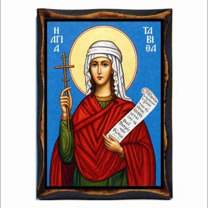 May include: A rectangular wooden plaque featuring a religious icon of Saint Tabitha. The figure wears a white head covering and a red cloak over a green garment, holding a thin cross and a scroll with Greek text against a bright blue background.