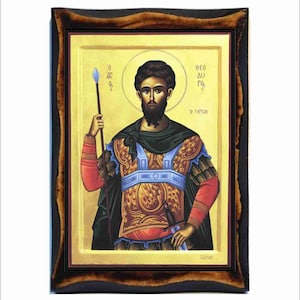 Pode incluir: Um ícone religioso que retrata uma figura masculina em armadura dourada e vermelha segurando uma lança e uma espada. O ícone tem um fundo dourado e uma moldura preta. O texto no ícone diz "ΘΕΟ ΔΟΞ Γ" e "Ο ΤΙΜΙΟΣ".