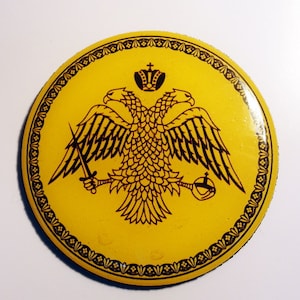 May include: A yellow round sticker with a black double-headed eagle design. The eagle has a crown on its head and is holding a sword and an orb. The sticker has a black border with a repeating pattern.