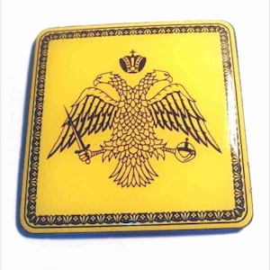 May include: A yellow square magnet with a black double-headed eagle design. The eagle has a crown on its head and is holding a sword and an orb. The magnet has a black border with a decorative pattern.