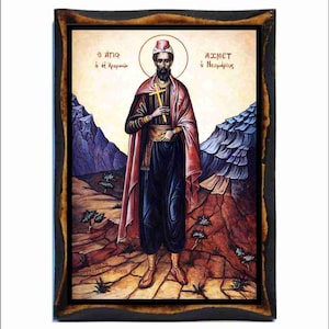 Puede incluir: Icono religioso enmarcado que representa a un hombre con un halo, sosteniendo una cruz. Viste un sombrero rojo, una túnica y una capa sobre ropa oscura. El fondo muestra un paisaje con montañas y árboles. El marco es marrón oscuro.