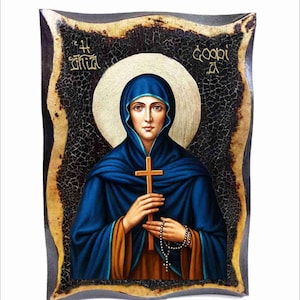 May include: An icon depicting a woman in a blue cloak holding a wooden cross and rosary beads. The image has a gold halo and is set against a dark, textured background with gold accents. The frame is dark with a gold border.