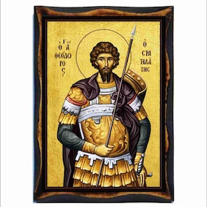 May include: Icon of a man in golden armor holding a spear, set against a gold background. The icon is framed in a dark, ornate wooden frame. The man has a halo and is depicted with a beard and dark hair. Greek text is visible on either side of the figure.