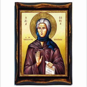 May include: An icon depicting a woman with a gold halo, wearing a blue headscarf and a dark purple robe. The icon is framed in a dark wood frame. The woman holds a scroll with text.