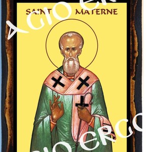 May include: A framed religious icon depicting Saint Materne, a Catholic saint. The icon features a yellow background with a green and pink robe, a white beard, and a black cross. The text "SAINT MATERNE" is written in black at the top of the icon.