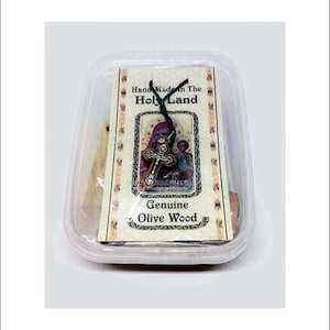 Puede incluir: Un pequeño recipiente de plástico transparente que contiene un collar de madera marrón en forma de cruz. El collar está en un cordón negro y tiene un pequeño dije de plata con una imagen de la Virgen María y el niño Jesús. El recipiente tiene una etiqueta blanca que dice "Hecho a mano en Tierra Santa" y "Madera de olivo genuina".