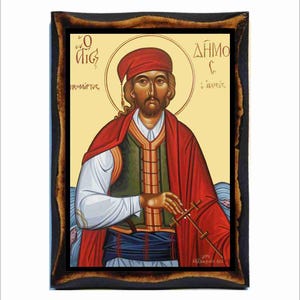 May include: A framed religious icon featuring a man in a red cap and cloak, holding a cross-like object. The icon has a gold halo and Greek text. The frame is dark brown with a textured appearance.