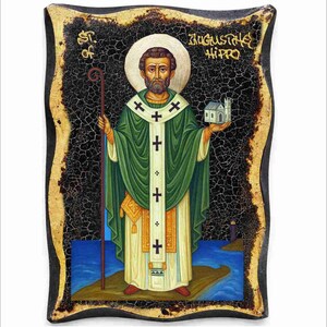 Op de afbeelding: Een religieus icoon met St. Augustinus van Hippo. Het kunstwerk toont een man in groene gewaden die een staf en een klein gebouw vasthoudt. De achtergrond is donker met gouden rand, en de tekst "St. of Augustine Hippo" is zichtbaar.