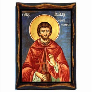 Puede incluir: Un icono pintado a mano de San Akakios, un santo ortodoxo griego. El icono está enmarcado en un marco de madera oscura y presenta un fondo azul con un halo amarillo alrededor de la cabeza del santo. El santo está representado con una túnica roja y sosteniendo una cruz. El texto "ΑΚΑΚΙΟΣ Ο ΘΕΟΣ ΚΑΙΜΑΚΙ" está escrito en griego en la parte superior del icono.