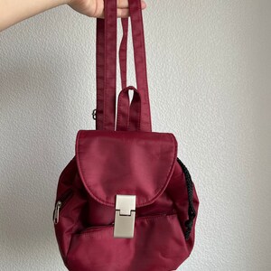 Puede incluir: Una mochila burdeos con cierre de solapa y cierre plateado. La pequeña mochila tiene un bolsillo frontal con cremallera y cierre de cordón negro. Las correas son burdeos.