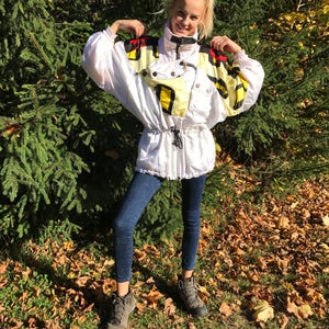 May include: A white vintage ski jacket with black, yellow, and red accents. The jacket has a high collar, a drawstring waist, and multiple pockets. The person is wearing dark blue jeans and gray hiking boots.