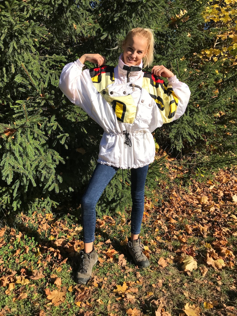 May include: A white vintage ski jacket with black, yellow, and red accents. The jacket has a high collar, a drawstring waist, and multiple pockets. The person is wearing dark blue jeans and gray hiking boots.