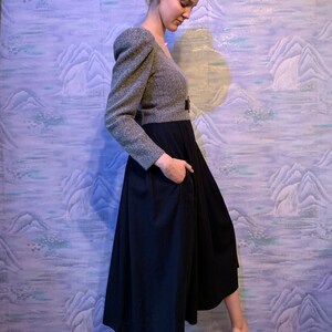 May include: A two-piece outfit featuring a grey tweed top with puffed sleeves and a black midi skirt. The skirt has a high waist and a flared silhouette. The outfit is accessorised with a black belt.