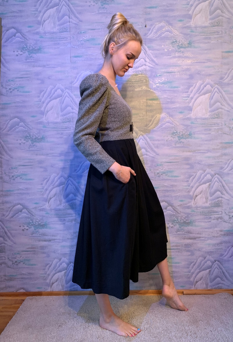 May include: A two-piece outfit featuring a grey tweed top with puffed sleeves and a black midi skirt. The skirt has a high waist and a flared silhouette. The outfit is accessorised with a black belt.