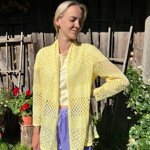 May include: A light yellow, open-front cardigan with a delicate, open-weave design. The cardigan has long sleeves and a patterned border. The person is wearing a light yellow top and purple pants. The background includes a wooden fence and greenery.