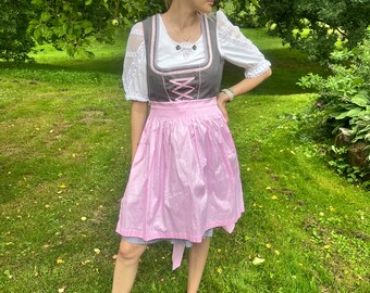 May include: A traditional German dirndl dress. The dress features a gray bodice with pink lacing, a white blouse with puffy sleeves, and a pink apron. The dress is worn over a blue and white checkered skirt.