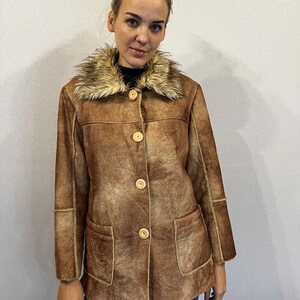 May include: A brown faux suede jacket featuring a tan faux fur collar, four light-colored circular buttons down the front, and two large patch pockets. The jacket has visible seam detailing along the sleeves and torso, providing a structured look.
