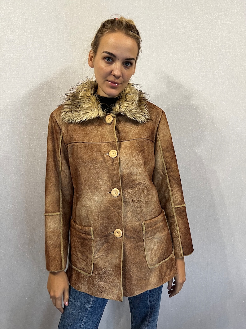 May include: A brown faux suede jacket featuring a tan faux fur collar, four light-colored circular buttons down the front, and two large patch pockets. The jacket has visible seam detailing along the sleeves and torso, providing a structured look.