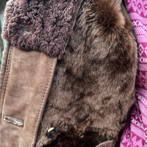 May include: Close-up of a brown shearling coat. The coat features a plush, dark brown fur lining and a suede-like exterior in a lighter brown shade. A button is visible, along with a portion of a patterned pink and white textile.
