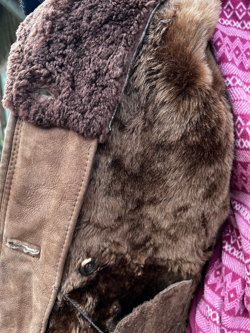 May include: Close-up of a brown shearling coat. The coat features a plush, dark brown fur lining and a suede-like exterior in a lighter brown shade. A button is visible, along with a portion of a patterned pink and white textile.