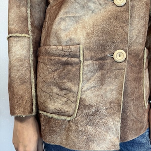 May include: A brown suede jacket with a visible shearling lining, featuring two large patch pockets on the front, exposed seams, and two light-colored circular buttons. The jacket is worn over blue denim jeans, showcasing a textured finish.