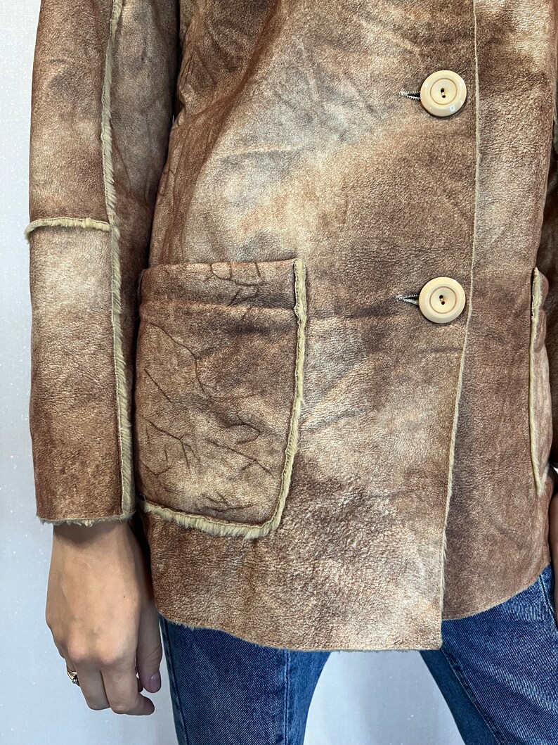 May include: A brown suede jacket with a visible shearling lining, featuring two large patch pockets on the front, exposed seams, and two light-colored circular buttons. The jacket is worn over blue denim jeans, showcasing a textured finish.