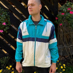 Könnte beinhalten: Eine türkisfarbene, weiße und marineblaue Windbreaker-Jacke mit Reißverschluss. Die Jacke hat eine Kapuze und ein Logo auf der linken Brust, das "Salomon" lautet.