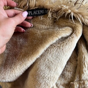 May include: A beige faux fur jacket with a long-haired collar and a soft, plush interior lining. A black rectangular tag is sewn into the neckline, featuring the white text PLACEMI and the letter M, indicating the size of the garment.