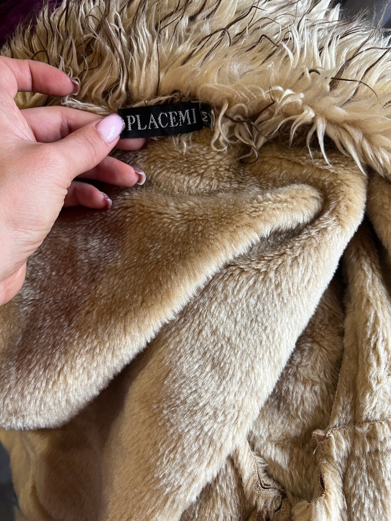 May include: A beige faux fur jacket with a long-haired collar and a soft, plush interior lining. A black rectangular tag is sewn into the neckline, featuring the white text PLACEMI and the letter M, indicating the size of the garment.