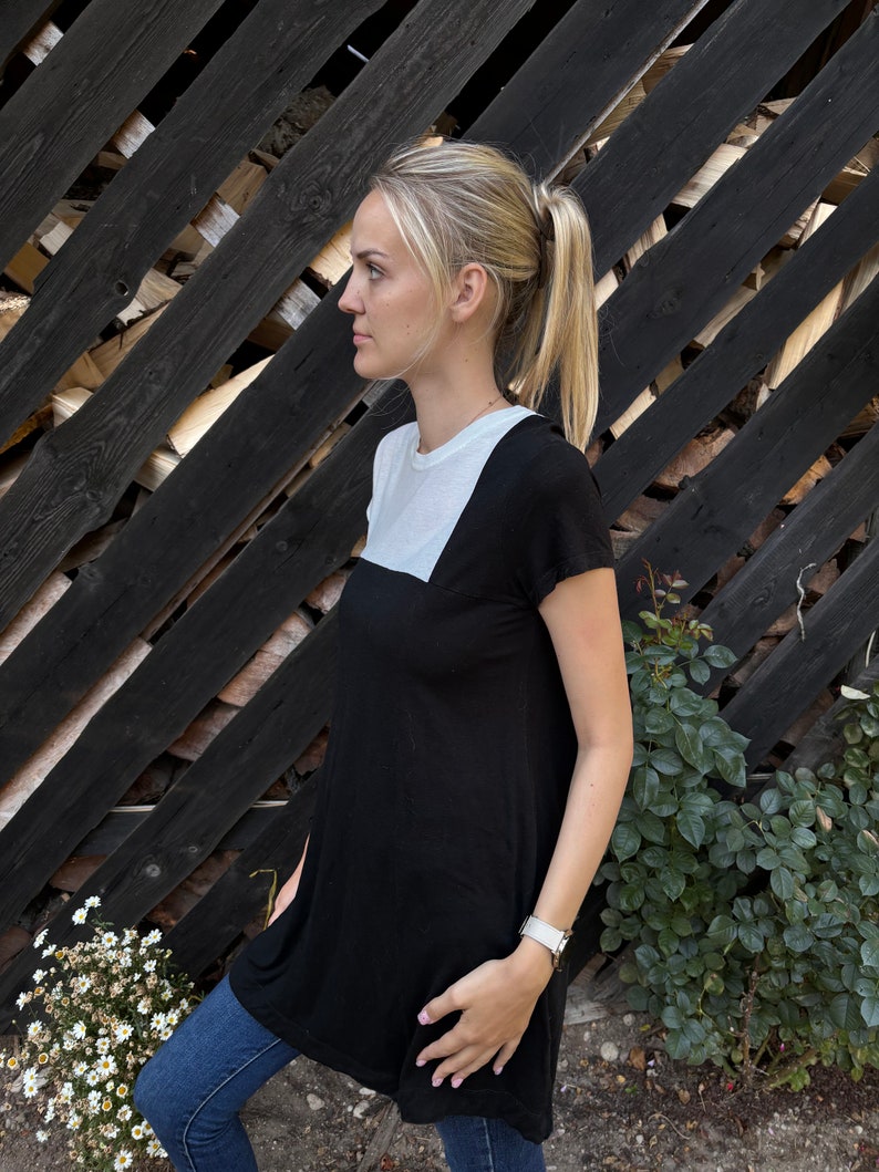 May include: A woman wearing a black and white tunic top with short sleeves. The top has a white panel across the chest and a black panel below. She is wearing blue jeans and a silver watch.