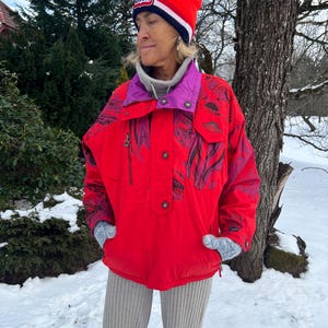 May include: A person wearing a red ski jacket with purple and black accents, a red beanie with a white stripe, and gray knit gloves. The person is standing in a snowy outdoor setting, wearing gray ribbed pants.