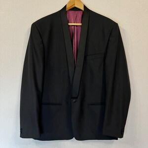 Puede incluir: Un blazer negro con solapa de satén y cierre de un botón, colgado de una percha de madera. El forro interior es de tela morada estampada. El blazer tiene dos bolsillos delanteros y botones en las mangas.