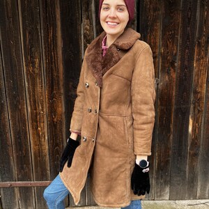 May include: A brown shearling coat with a double-breasted design. The coat features a sheepskin lining and a wide collar. The person in the image is wearing black gloves, blue jeans, and a burgundy headband. The background is a wooden door.